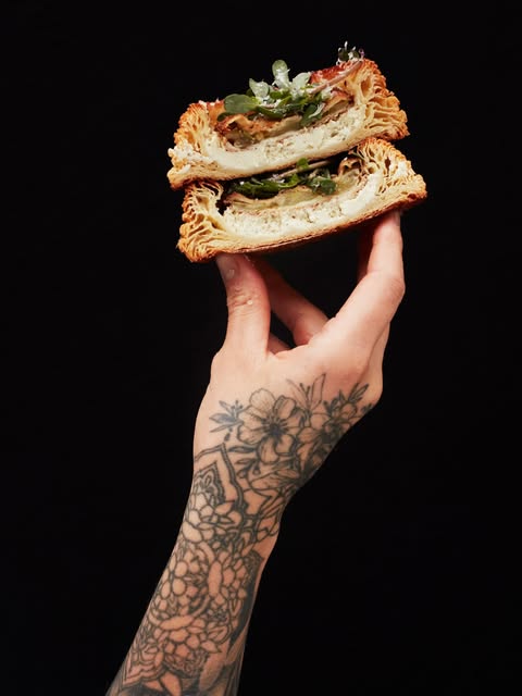 A tattooed hand holding two halves of a Black Magic Bakehouse croissant sandwich